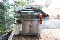 Full view of stainless steel Char-Broil infrared BBQ on wooden deck, with propane tank at left and boxed rotisserie kit on right shelf under grill cover