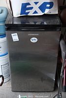 Front view of Sylvania mini fridge with stainless steel door and black top showing some surface wear, closed door with manufacturer badge and a promotional sticker.