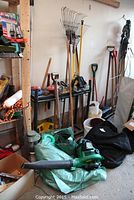 Overall view showing leaf blower, rakes, shovels, tarp, and other garden items leaning against a wall in a garage or shed.