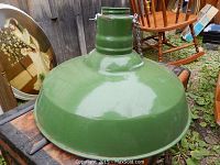 View of a large green enamel industrial lamp shade resting on a wooden crate with a rocking chair in the background, showing the rounded design and glossy enamel finish.