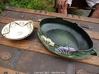 Roseville 466 oval pottery dish with green matte finish and raised floral decoration, alongside Noritake divided candy dish with floral painting and gold trim, placed on wooden surface outdoors.