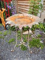 Full side view of the metal sunflower bird bath outdoors on gravel with flowers and plants nearby