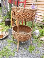Full view of the two-tier woven basket plant stand with detailed weaving on the top basket and plain weaving on the bottom basket standing on gravel with greenery around.