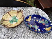 Both Majolica dishes shown side by side on a wicker surface, one yellowish with handles, the other dark blue with colorful raised patterns.