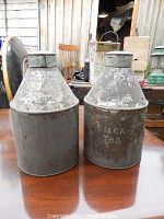 Photo of two tall, conical vintage metal tea tins placed on a wooden table. Both have heavily weathered surfaces, one with faded 'Black Tea' lettering.