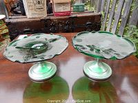 Pair of green glass pedestal candy dishes with scalloped edges, shown on wooden table, photographed outdoors.
