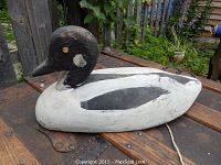 Side profile of the duck decoy showing black head with yellow eyes, white cheek patch, and white body with black wing patches lying on a rusty metal surface outside.