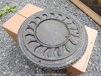 Cast iron floor grate placed outdoors on a cardboard box, showing the circular design with concentric rings and patterned openings.