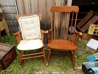 Two older wooden rocking chairs shown side by side outdoors on grass with a wooden wall background. One chair has a tufted cushion and pressed back design. The other chair has vertical slats and a repair on the side.