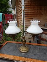 Front view of antique brass student lamp with two white milk glass shades on tray outdoors against wooden siding and barrel backdrop.