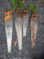 Four vintage hand saws laid out on gravel with visible wear, rust, and wooden handles.