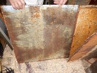 Photo showing two rusted square metal ceiling tins with circular embossing, held upright by hands, surface shows rust and wear.