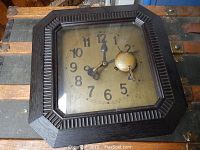 Square 1930s German wood case wall clock with carved ridged detailing along the inner frame, showing a worn golden face and black Arabic numerals, and a key winding mechanism attached.