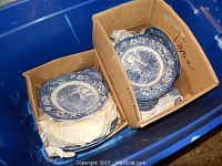 Two cardboard boxes containing multiple blue and white decorative plates with floral borders and American scenes, in blue plastic bin.