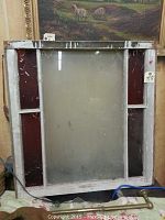 Front view of the antique stained glass window showing central clear pane and red stained glass side panels in a worn white wooden frame.