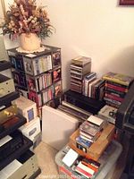 Wide view showing black metal CD rack filled with jewel-case CDs, LP records in sleeves, cardboard box, plastic bin of tapes and adjacent electronics stack