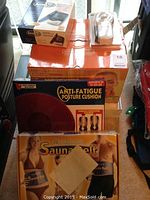 Photo of stacked healthcare items including Smartmask Home Spa Treatment box, Anti-Fatigue Posture Cushion box, and Velform Saunabelt box showing front packaging and partial view of individual items