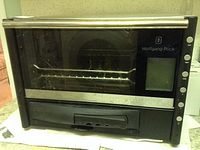 Front view of Wolfgang Puck black and stainless steel toaster oven showing glass door, crumb tray closed, and control panel on the right side.