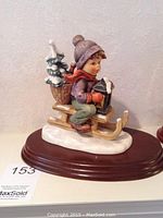 Front side of Goebel Hummel Ride into Christmas figurine showing the child on sled holding a lantern with basket of snowy trees on back