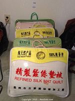 Four refined silk mat quilts stacked in their storage bags, two of which are clearly labeled with '100% pure natural silk' and 'Refined Silk Mat Quilt' in English and Chinese.
