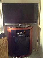Full view of 40" Sony Bravia LCD TV on wooden swivel stand cabinet with glass door; remote on top shelf.
