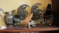 Five metal decorative pieces displayed on a wooden shelf including a pitcher, two rooster sculptures, a grand piano sculpture, and a person playing piano sculpture.