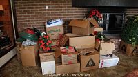 Wide view showing multiple cardboard boxes containing various Christmas decorations stacked near fireplace and wall, including tree parts and assorted decorations.