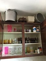 Upper kitchen cabinet showing aluminum pots on top and multiple shelves filled with various glasses and containers.