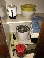 Cabinet shelf containing white electric coffee maker with glass carafe, plastic mixing bowls (yellow and white), ice cube tray with blue lid, white appliance base possibly food processor, red tin container, and white kitchen scale with dial.