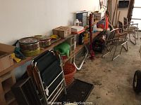 View of wooden shelf against wall on left side showing folding chairs, metal tins, boxes, tools, basket and plastic bucket on concrete floor.