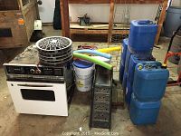 Overall view showing a white GE oven with stacked metal vehicle ramps, circular metal hub caps on top, foam pool noodles, white buckets, and multiple blue plastic gas cans.