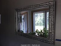 Large rectangular mirror on wall reflecting room interior, framed with textured silver and black checkered pattern.