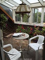 Patio area with white plastic chairs and a round metal and glass top table. Hanging stained glass lamp above table, surrounded by potted flowering plants.