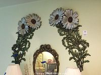 Two tall and large metal floral wall sculptures with big daisies and leaves, photographed on wall above decorative mirror and lamps.