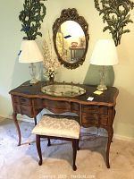 Full view of wooden vanity desk with drawers, two lamps on top, matching stool and oval mirror overhead