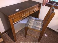 Side angle photo showing one wooden desk with two drawers and one matching wooden chair with plaid seat. Desk's surface is dark brown with lighter wood frame.