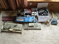 Overview of multiple computer keyboards, a flexible keyboard in box, PC cooling fans, power adapters, and a desk lamp laid out on carpeted floor.