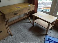 Photo of wooden desk with two drawers, angled wood legs, and metal handles showing surface wear and scratches alongside a small rolling wooden table with lower shelf and caster wheels.