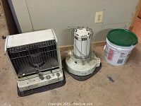 Two kerosene heaters side by side on floor near wall and electrical outlet