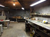 Wide view of workroom showing wooden workbenches, shelving, various materials, and exposed ceiling.