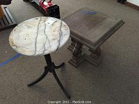 Both end tables side by side, the plastic one is square with scratches, the wood and marble topped table is square with decorative legs and a round white marble top.