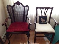 Two wooden chairs side by side, the larger wing-backed one with red velvet cushion and cane backrest; smaller chair with white cushion and dog pillow.