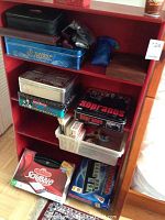 Red shelf with various board and card games. Visible games include Monopoly, Sopranos, Alberta Fishing Derby, and Scrabble box set outside of lot. Also includes black case and a plastic container with many card decks.