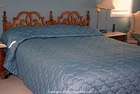 Wooden bed with blue quilted comforter spread over mattress, decorative wood headboard visible, bedside table and lamp partially visible.