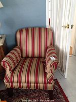 Full front view of plush upholstered armchair with red and gold vertical stripes with blue accents, shown in room on patterned carpet.