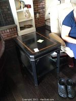 Full view of black wooden side table with inset border and lower shelf next to beige armchair, showing overall style and dimensions.