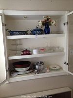 Upper cabinet shelf with blue floral enamel serving dishes, blue glass vase with flowers, glass display trays, red glass covered dish, white ceramic dishes, glass measuring cup, small clear glass bowls, large salad bowls, metal pots and kitchen utensils.