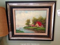 Framed oil on canvas painting showing a cottage with a red roof by water with trees and a sailboat in the distance.
