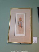 Framed art print of a girl on a bicycle, vertical orientation, gold frame and beige mat, hanging on wall.