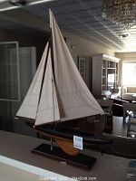 Full side view of the model sailboat showing the cream canvas sails, black hull, and wooden base stand in a room setting.